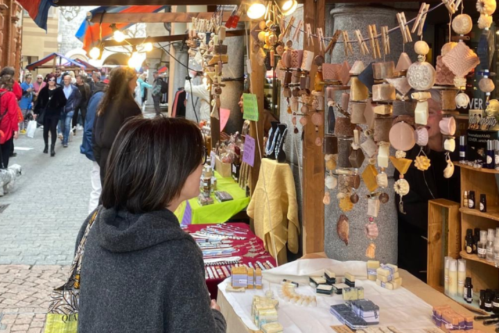 Person browsing soap and ornaments at outdoor market stall, with people walking in background.