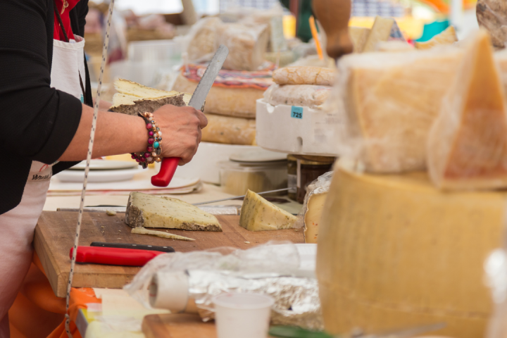 Person cutting cheese at a market stall with various cheese wheels displayed.