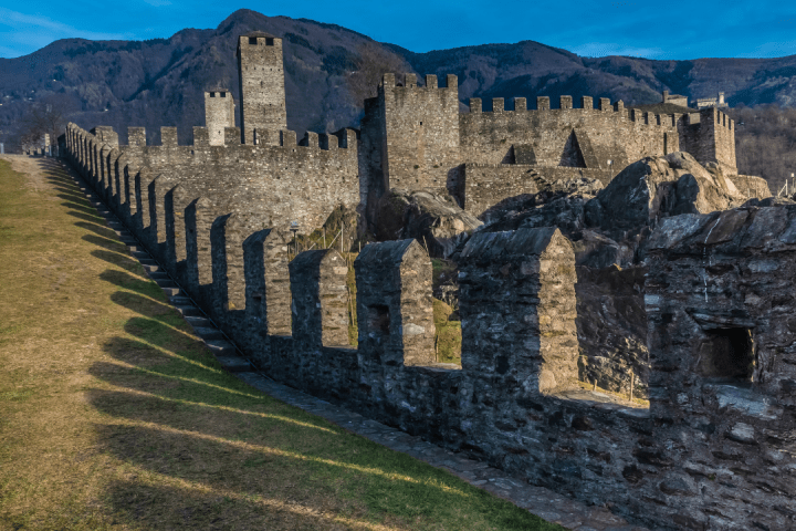 Stone castle with walls and towers against mountain backdrop under blue sky.