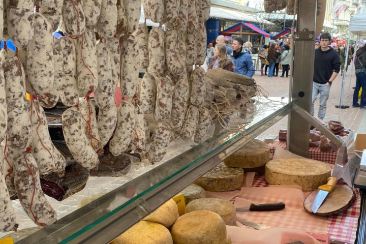 Market stall with hanging sausages and cheese display, people browsing in background.
