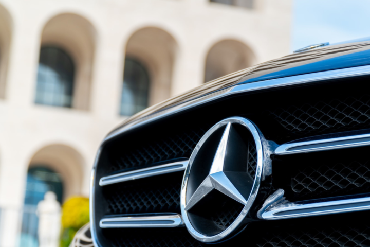 Close-up of a car grille with Mercedes-Benz logo, arches of a building in the background.