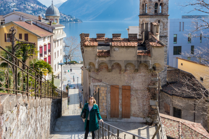 Woman walking up stairs in a scenic European town, with mountains and lake in the background.