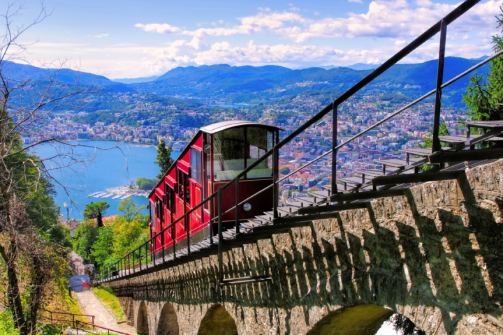 Red funicular on a steep hill with a view of a lake and city below.