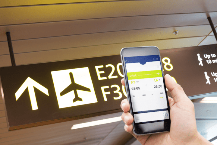 Hand holds phone with flight info, airport signs above show gate directions and walking times.