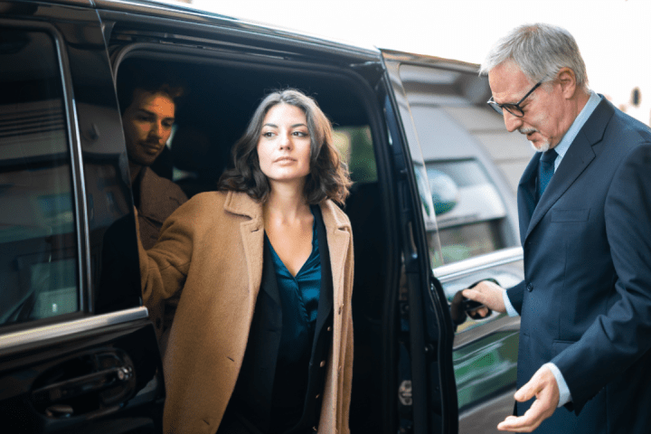 Woman exits a black car assisted by a suited man with another man inside.