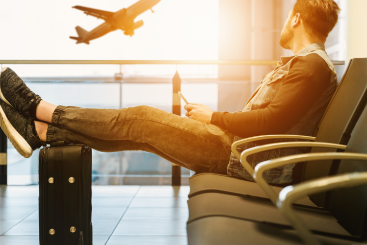 Person sitting in airport lounge, feet on luggage, watching plane take off.